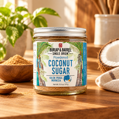 A jar of Burlap &amp; Barrel Powdered Coconut Sugar sits on a wooden table, surrounded by a halved coconut, bowls of the brown sugar, and a green plant in a warm, natural setting.