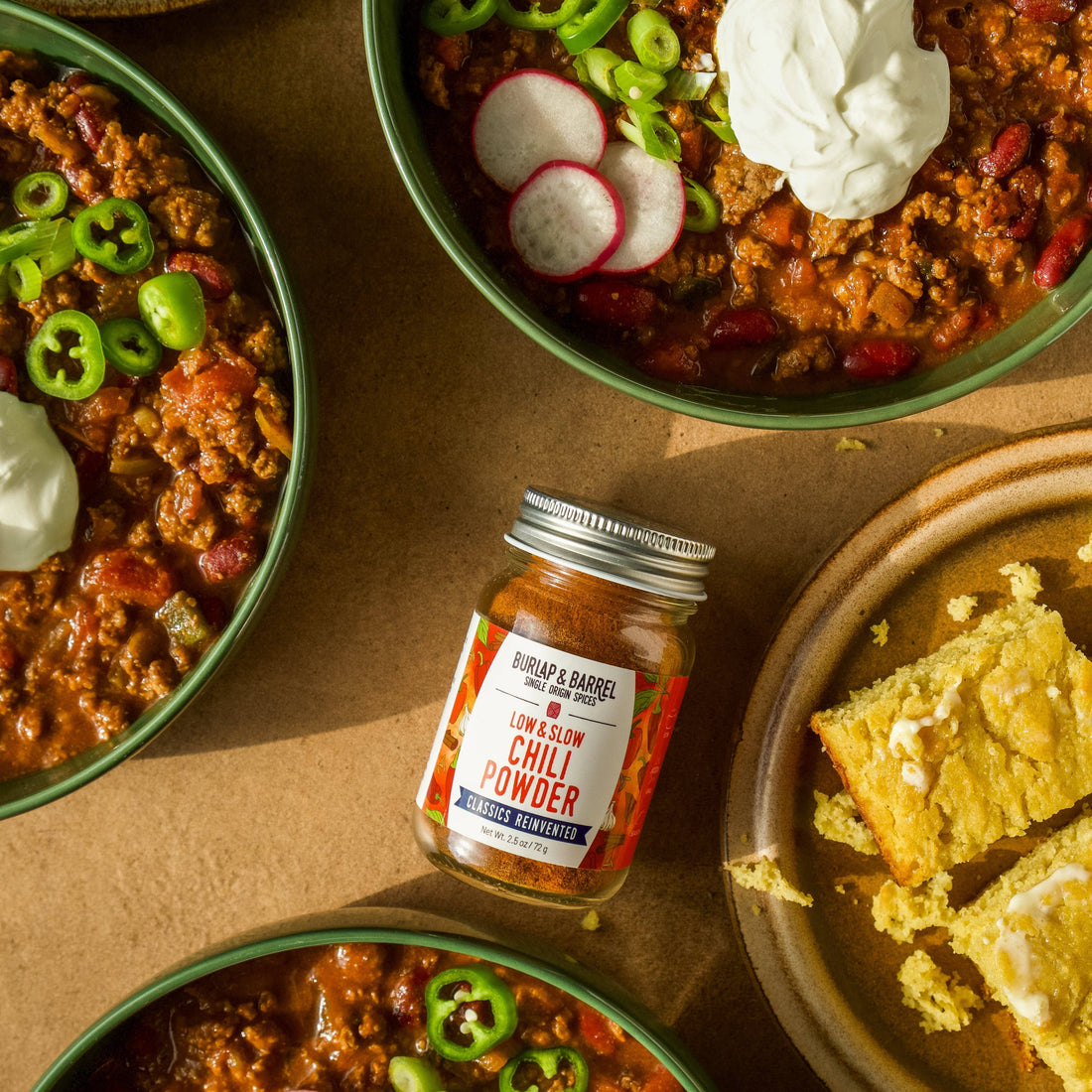 Bowl of chili with cornbread and a jar of chili powder on a brown surface