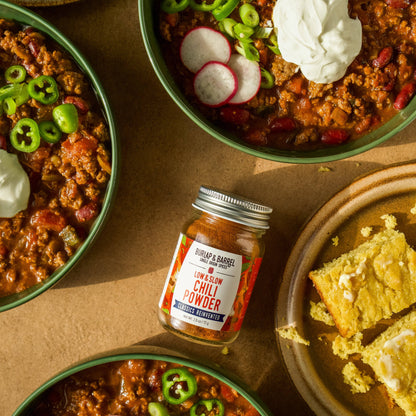 Bowl of chili with cornbread and a jar of chili powder on a brown surface