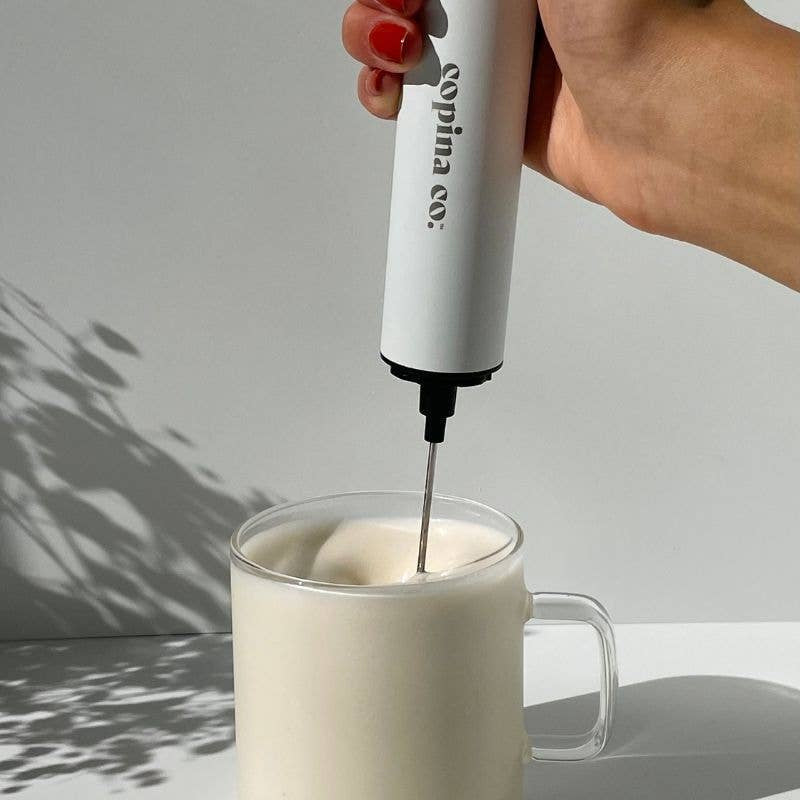 Hand using a white milk frother to create foam in a clear mug on a light gray background.