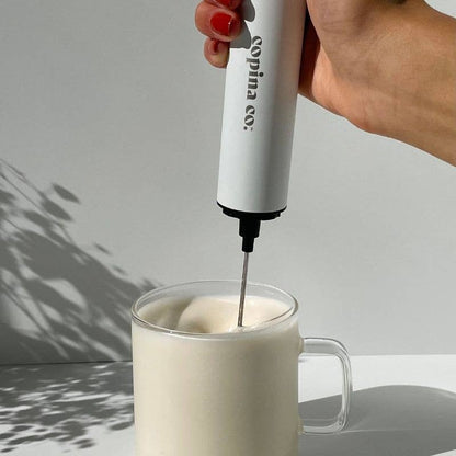 Hand using a white milk frother to create foam in a clear mug on a light gray background.