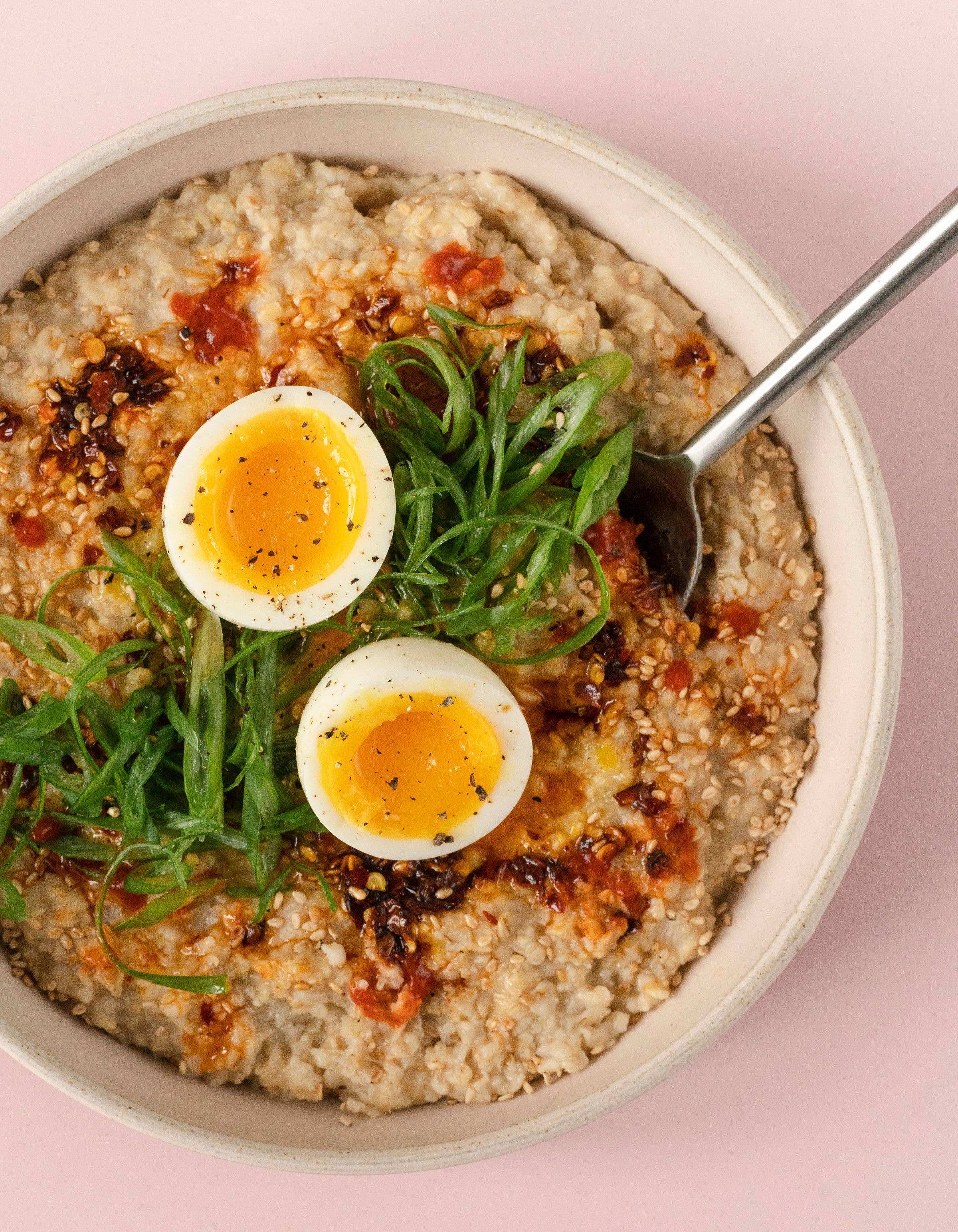 Bowl of savory porridge with soft-boiled eggs and greens on a pink background
