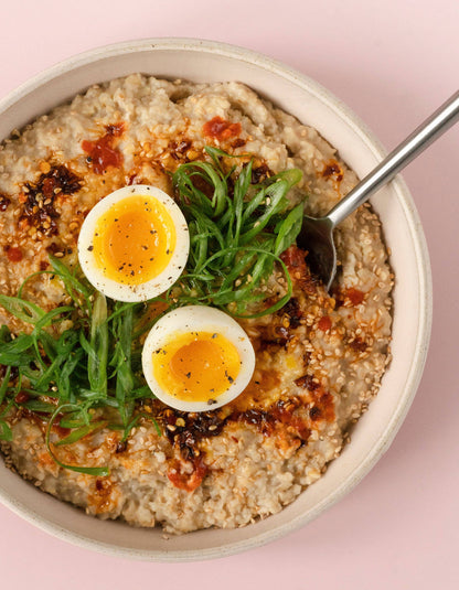 Bowl of savory porridge with soft-boiled eggs and greens on a pink background