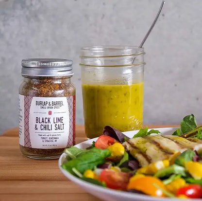 A jar of Burlap &amp; Barrel Black Lime &amp; Chili Salt stands on a wooden table beside a glass jar of yellow vinaigrette dressing and a salad plate in the foreground.