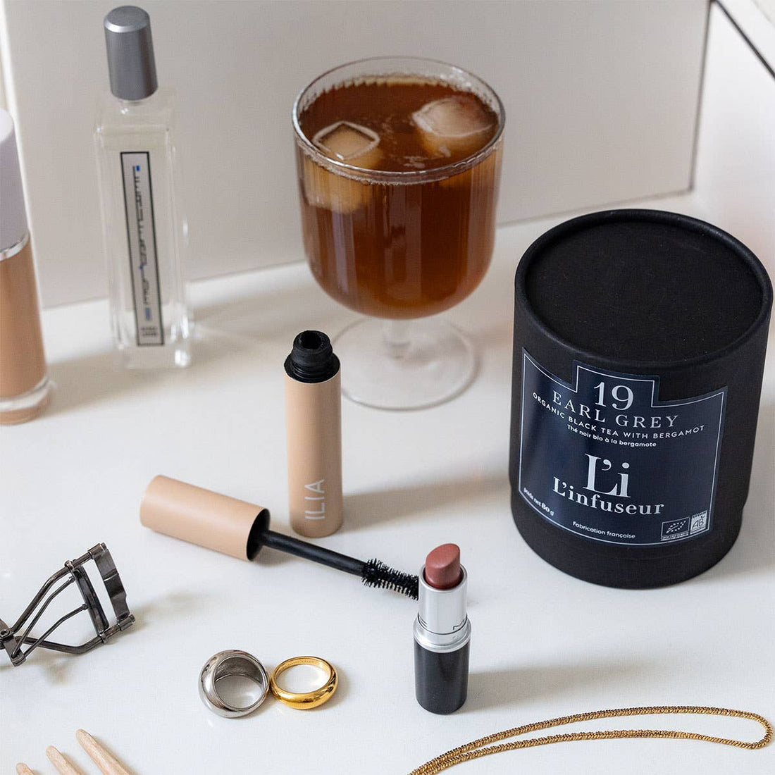 Makeup items and a candle on a white surface with a glass of iced tea in the background.