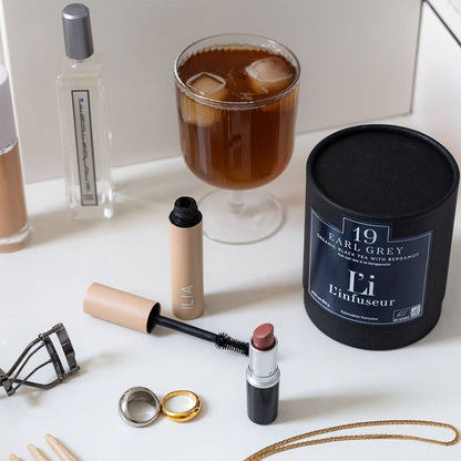 Makeup items and a candle on a white surface with a glass of iced tea in the background.