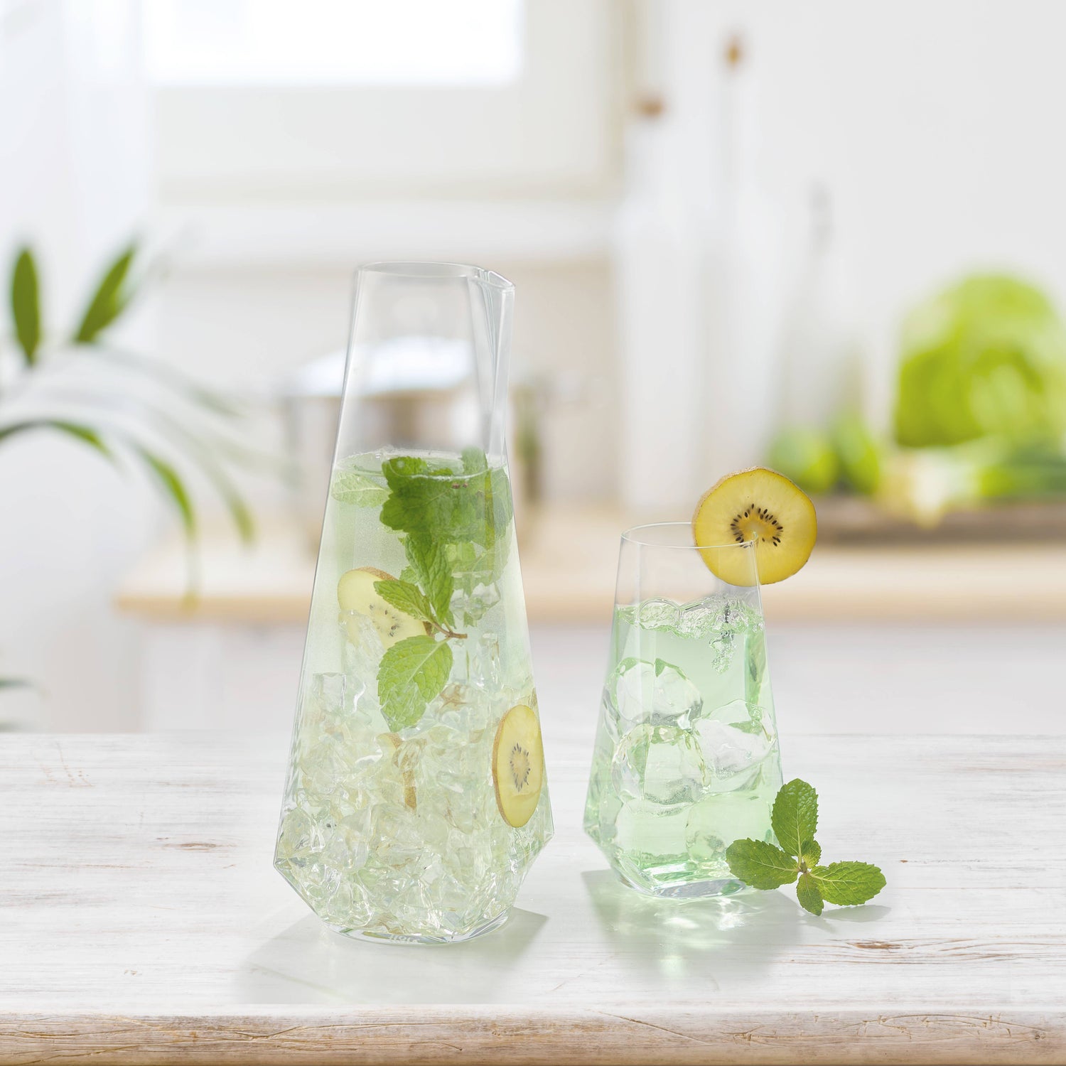 Clear carafe and glass with green liquid, kiwi slices, and mint leaves on a light surface.
