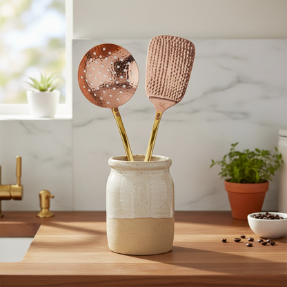 A skimmer and spatula with hammered copper-colored heads and gold handles, standing in a ceramic jar on a wooden countertop in a bright kitchen setting.