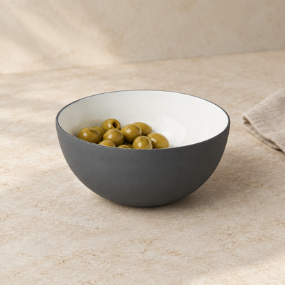 A dark gray and white bowl filled with green olives, set on a textured beige stone surface with a neutral linen napkin in the background.
