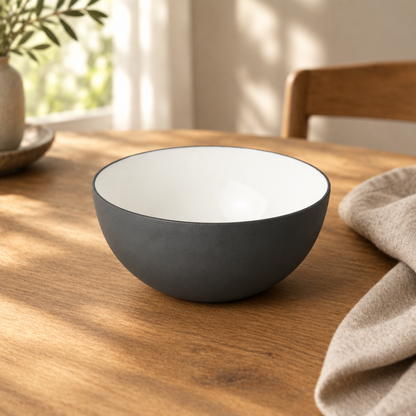 A dark gray and white bowl sits on a wooden dining table in a warm, sunlit room with a chair and plant in the background.
