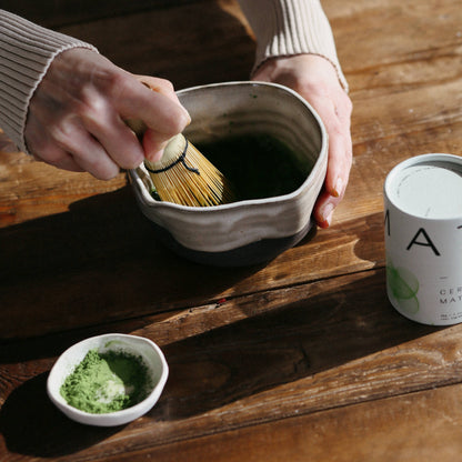 Person preparing matcha tea on a wooden table with a canister labeled &