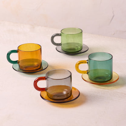 Four translucent colored glass mugs with matching saucers arranged on a light beige surface. The mugs come in amber, green, teal, and smoky gray, each with a different colored handle.