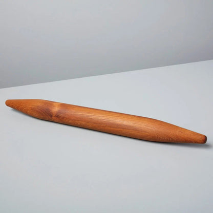 A single teak wood rolling pin with a smooth, polished finish and visible wood grain, resting diagonally on a light gray surface against a neutral gray background.